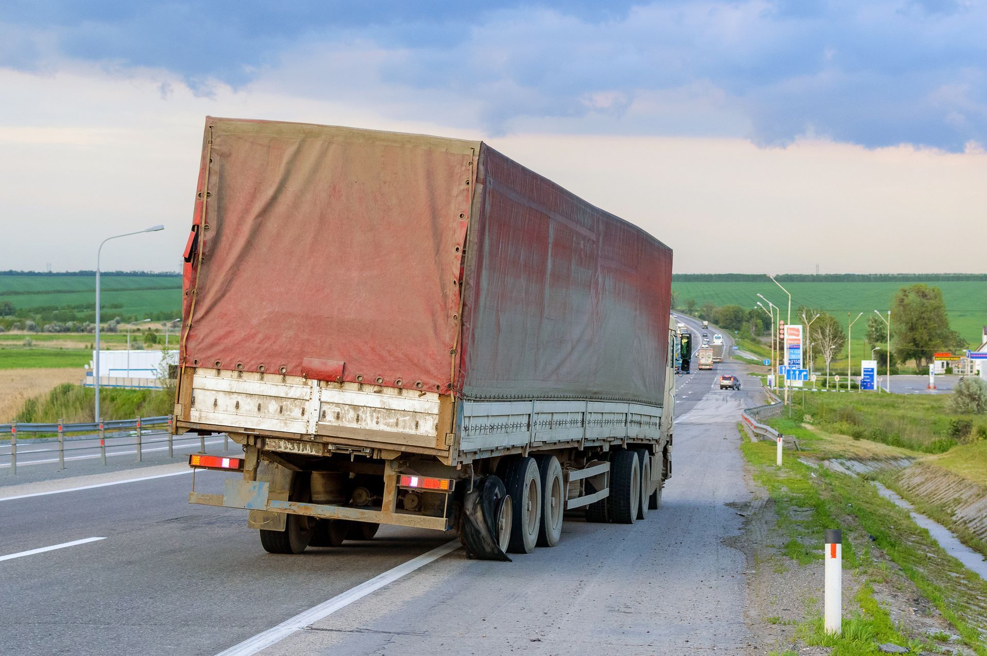 Large semi-truck with red tarp travels on a highway, with green fields and cloudy sky in the background.