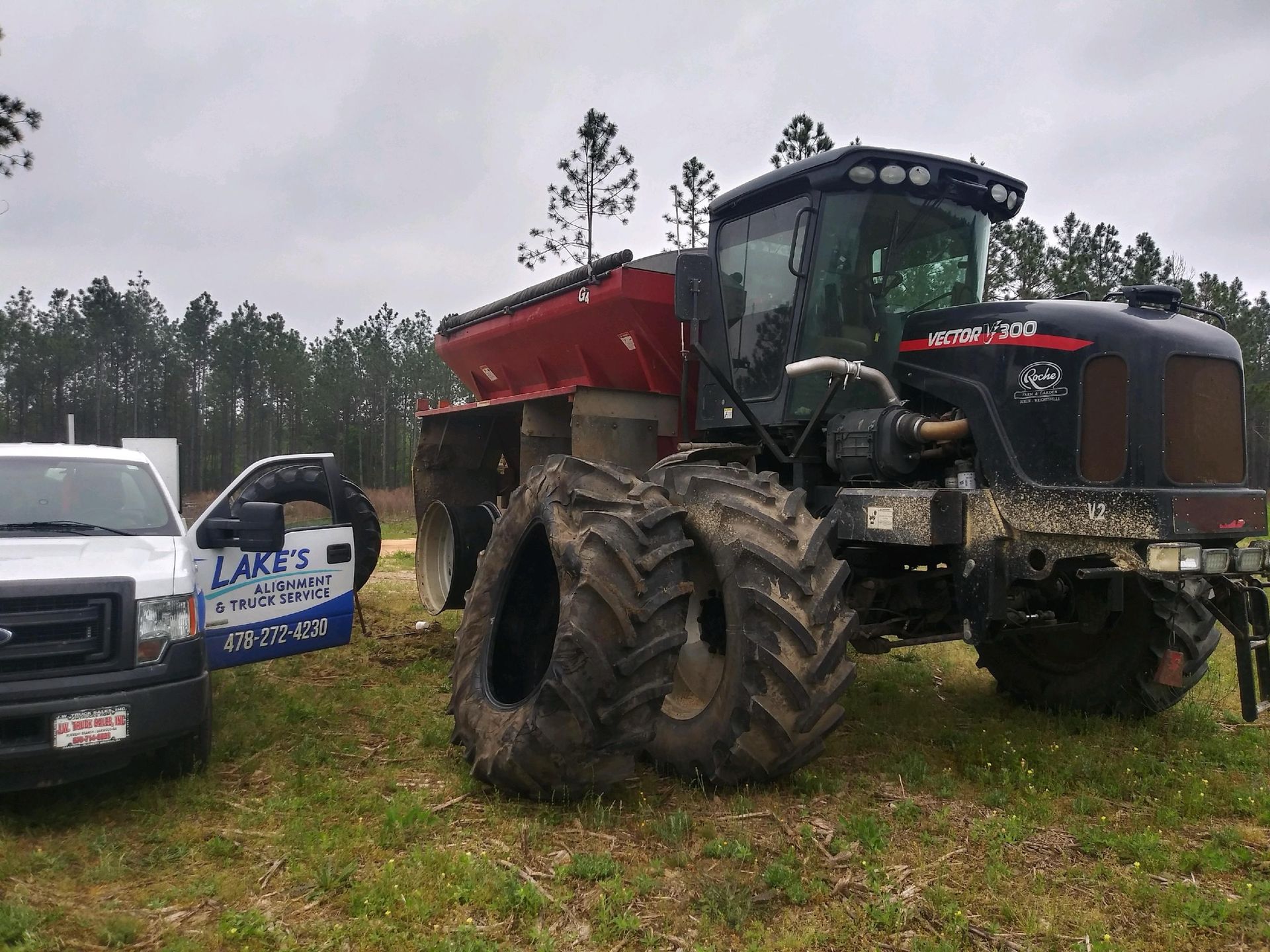 Black tractor with red hopper and mud-caked tires sits near a white truck in a field.