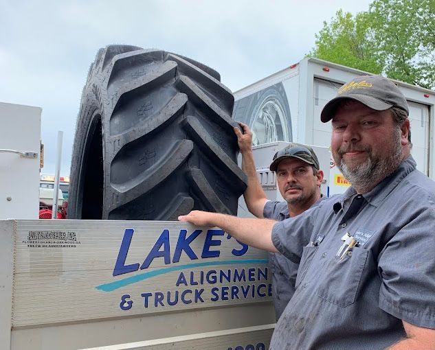 Two men stand with a massive tractor tire on a truck bed. The truck says "Lake's Alignment & Truck Service".