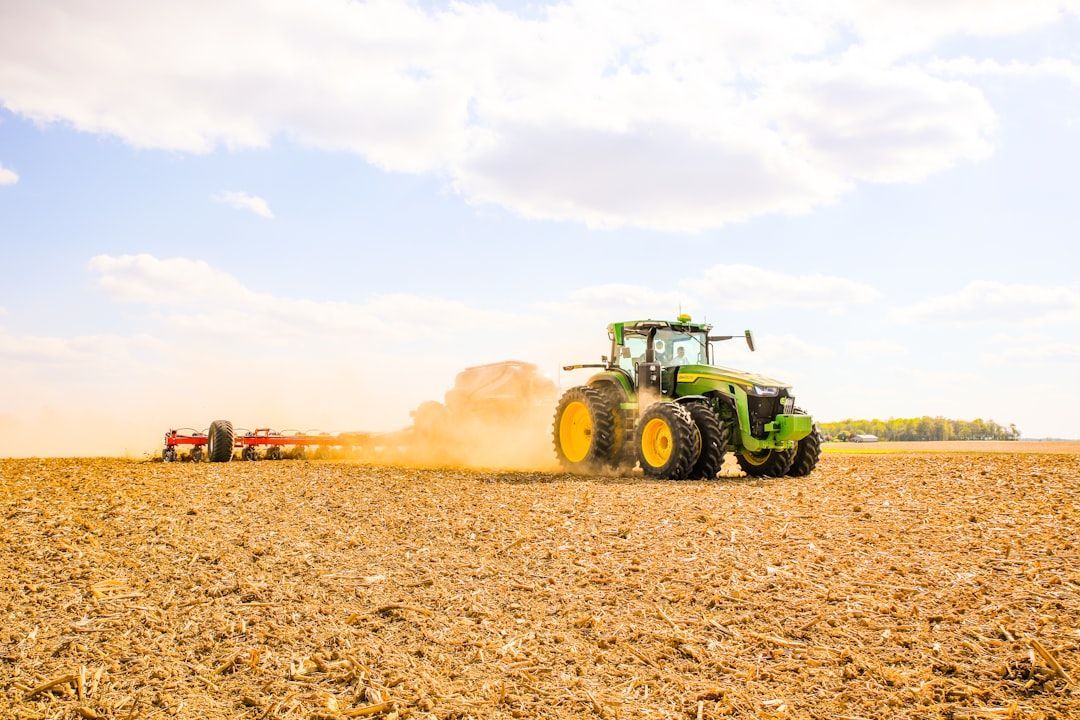 Green tractor plowing a field, kicking up dust under a cloudy blue sky.