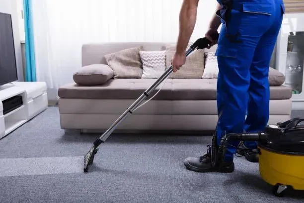 Cleaner vacuuming a gray carpet beside a beige sofa in a living room