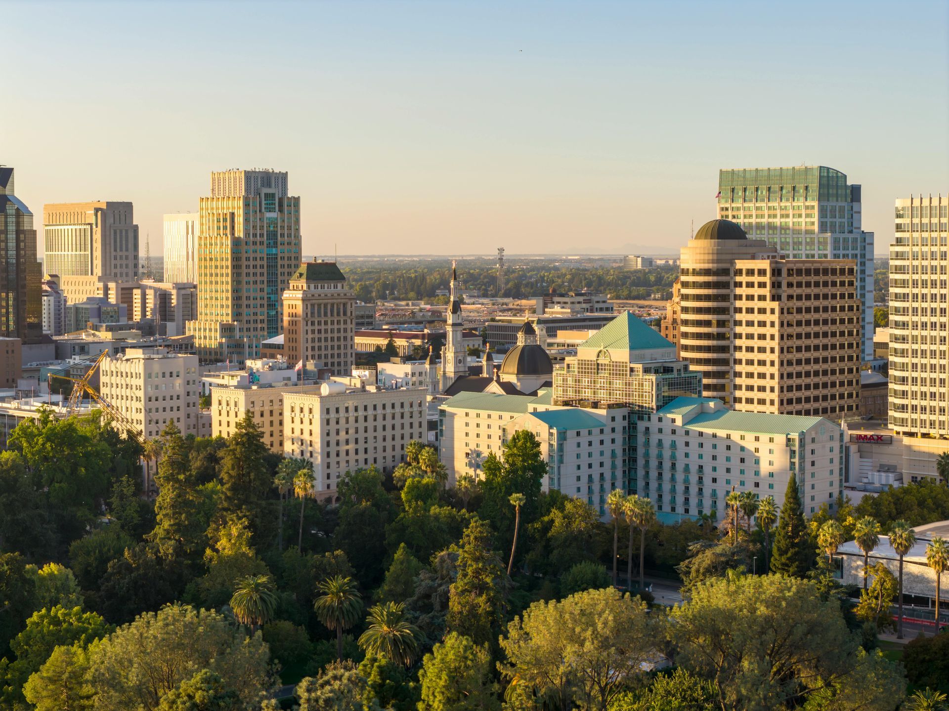 City skyline at sunset, with buildings of various heights and green trees in the foreground.