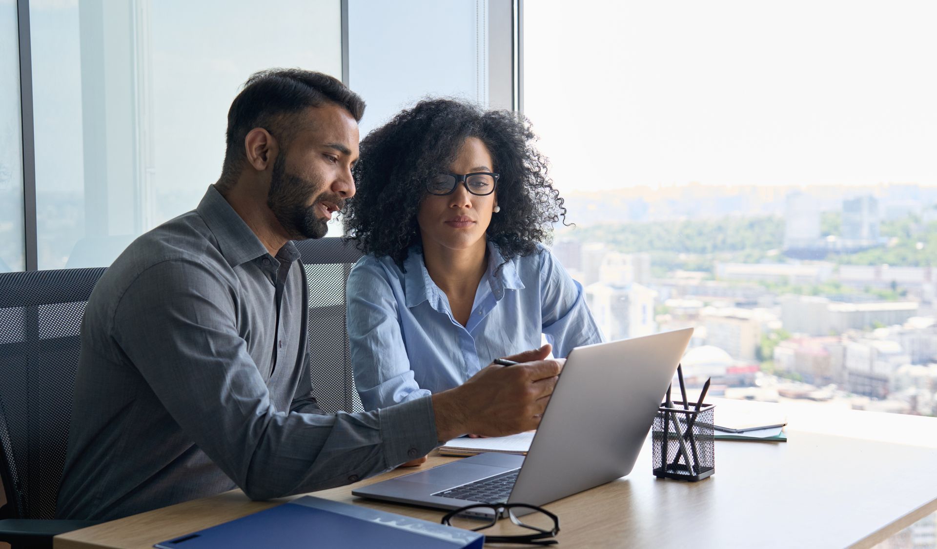 Man points at laptop screen, showing a woman, both in an office, city view in the background.