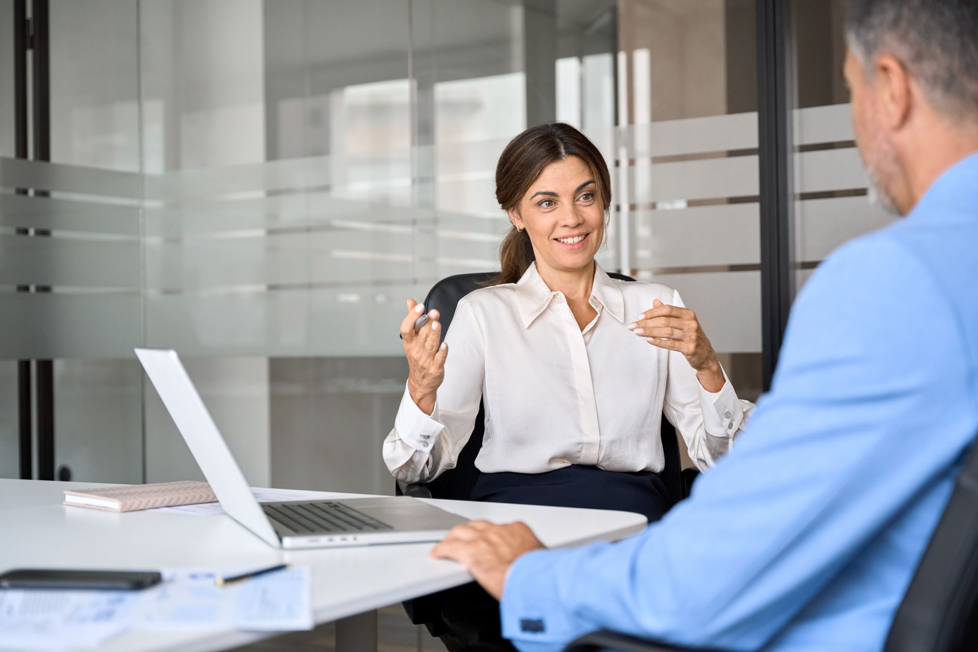 Woman in a white blouse gestures while speaking to a man in a blue suit at an office desk with a laptop.