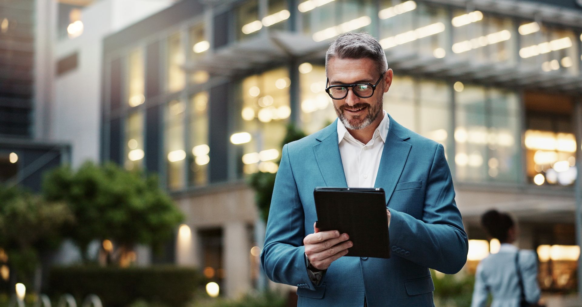 Man in suit with glasses looking at a tablet in front of a modern building.