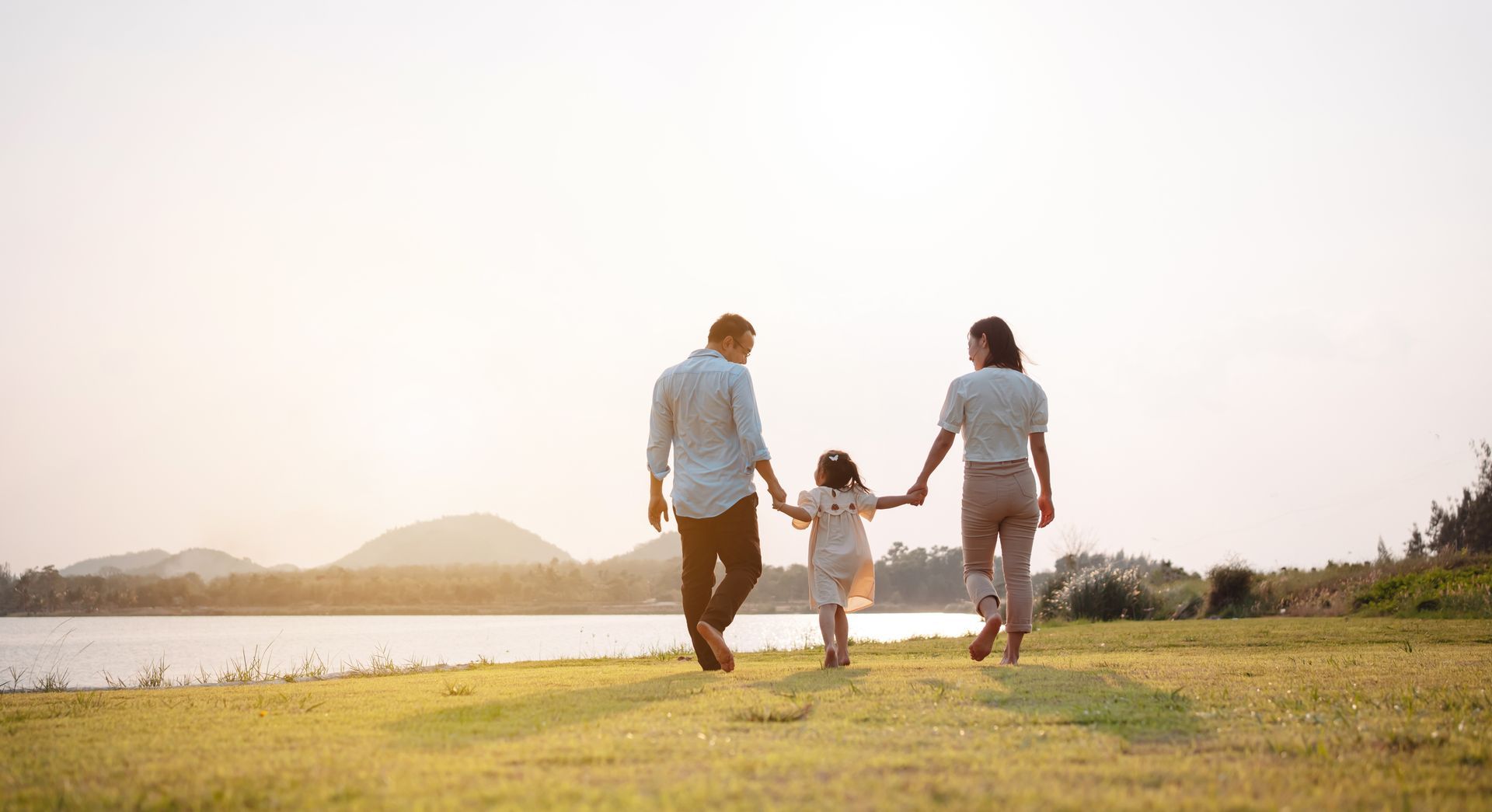 Family of three walking hand-in-hand along a lake at sunset.