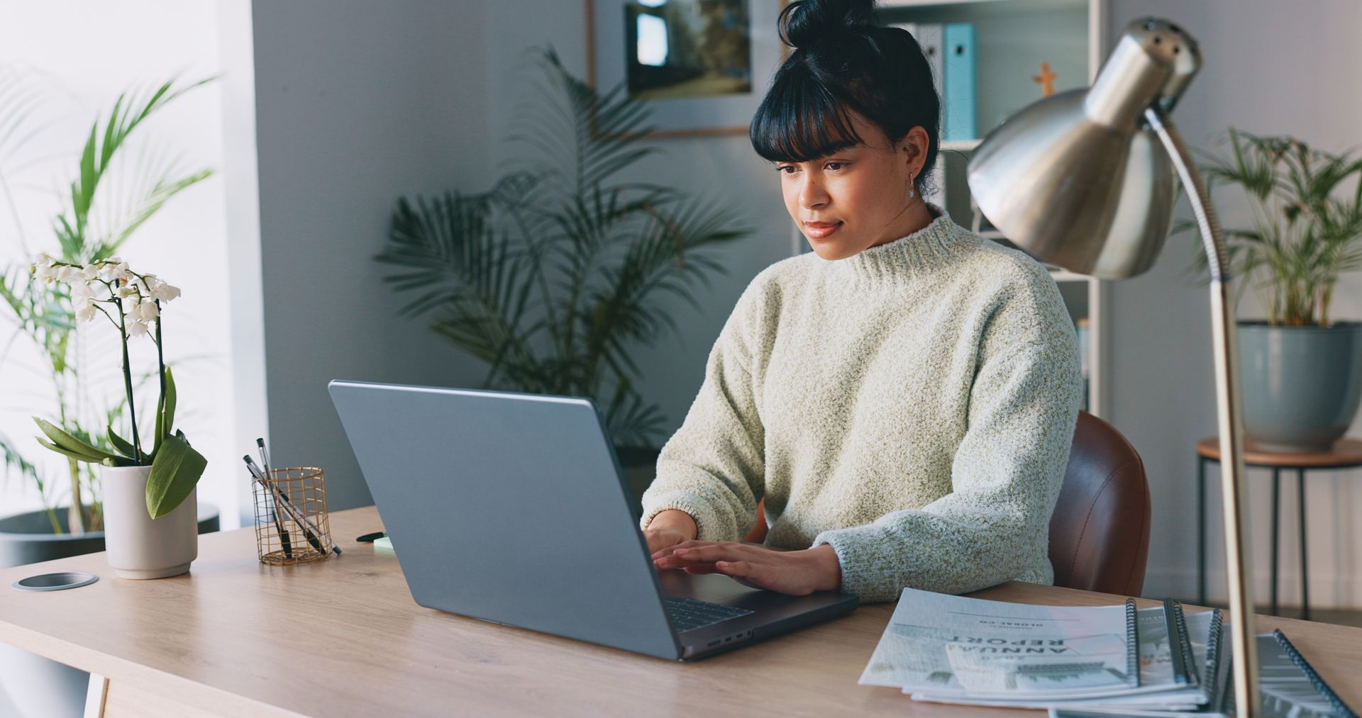 Woman working on laptop at desk, plants and lamp in home office.