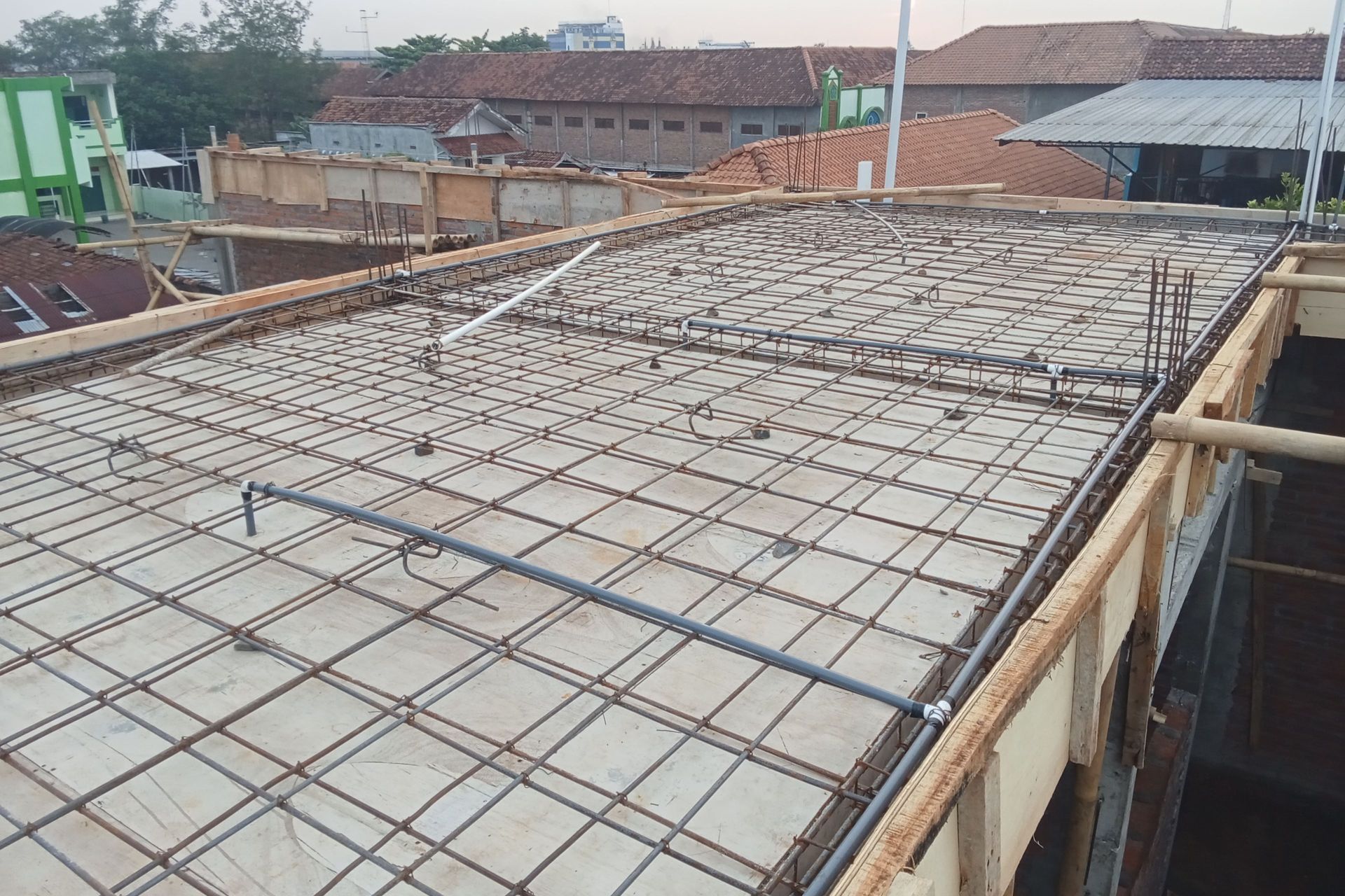 Construction site: reinforced steel grid for a concrete slab, with pipes laid, wooden framing, and building exteriors in the background.