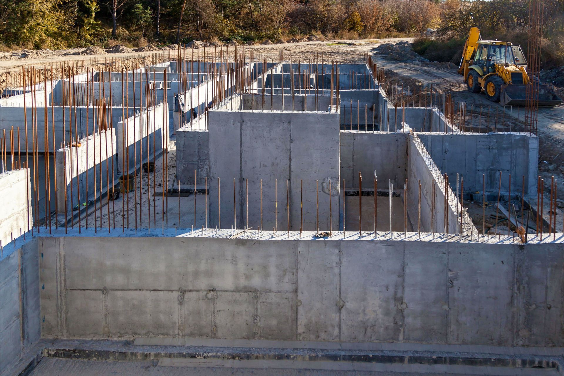Concrete foundation for a building under construction, with rebar sticking out and a backhoe in the background.