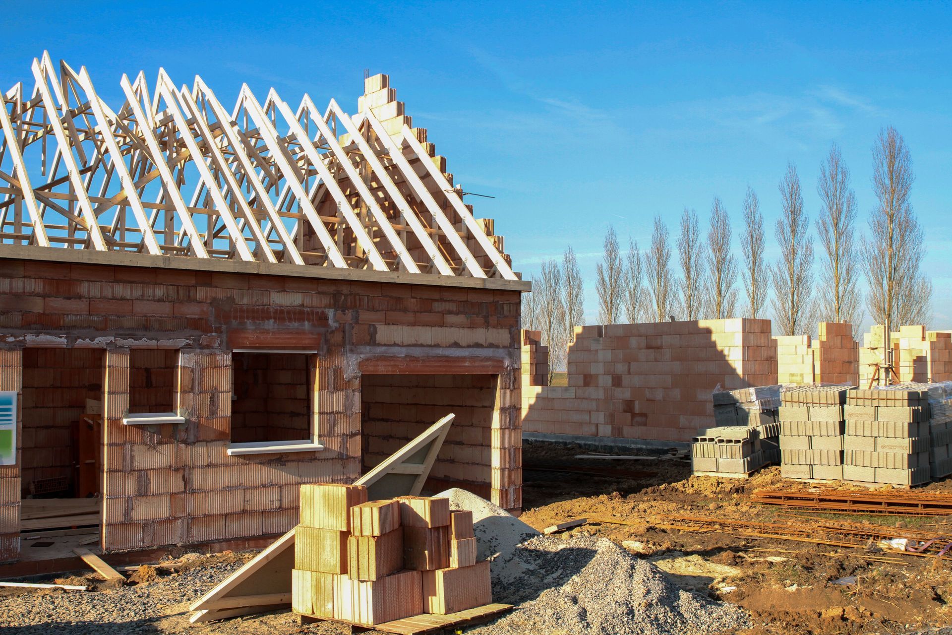 Construction site with brick structures, wooden roof framing, and stacks of bricks on a sunny day.