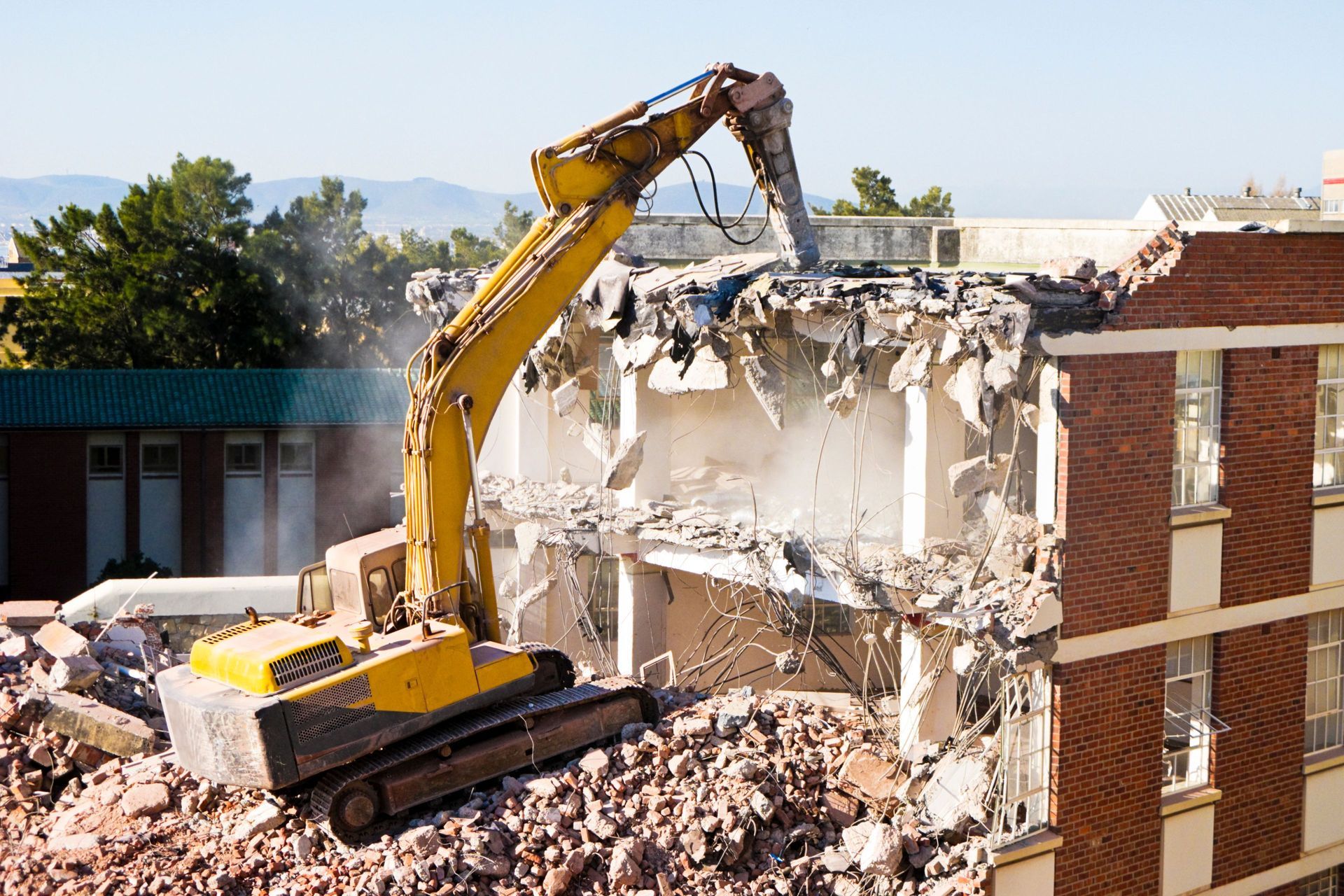 A yellow excavator demolishes a brick building on a sunny day. 