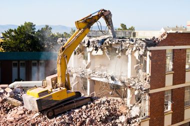 A yellow excavator demolishes a brick building, rubble piles below.
