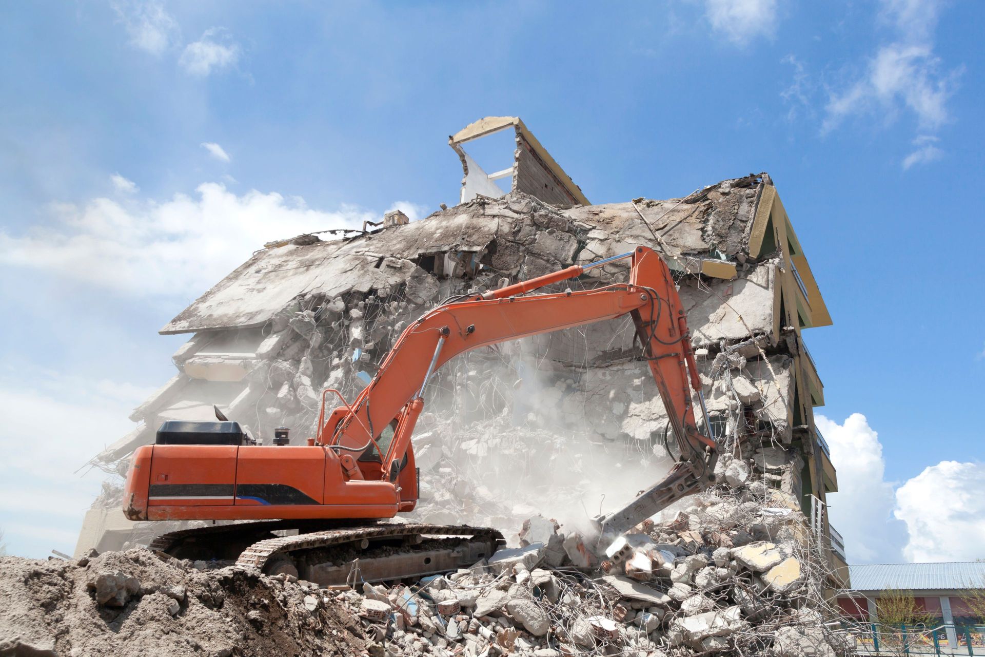 Orange excavator demolishing a building against a blue sky, debris in foreground.