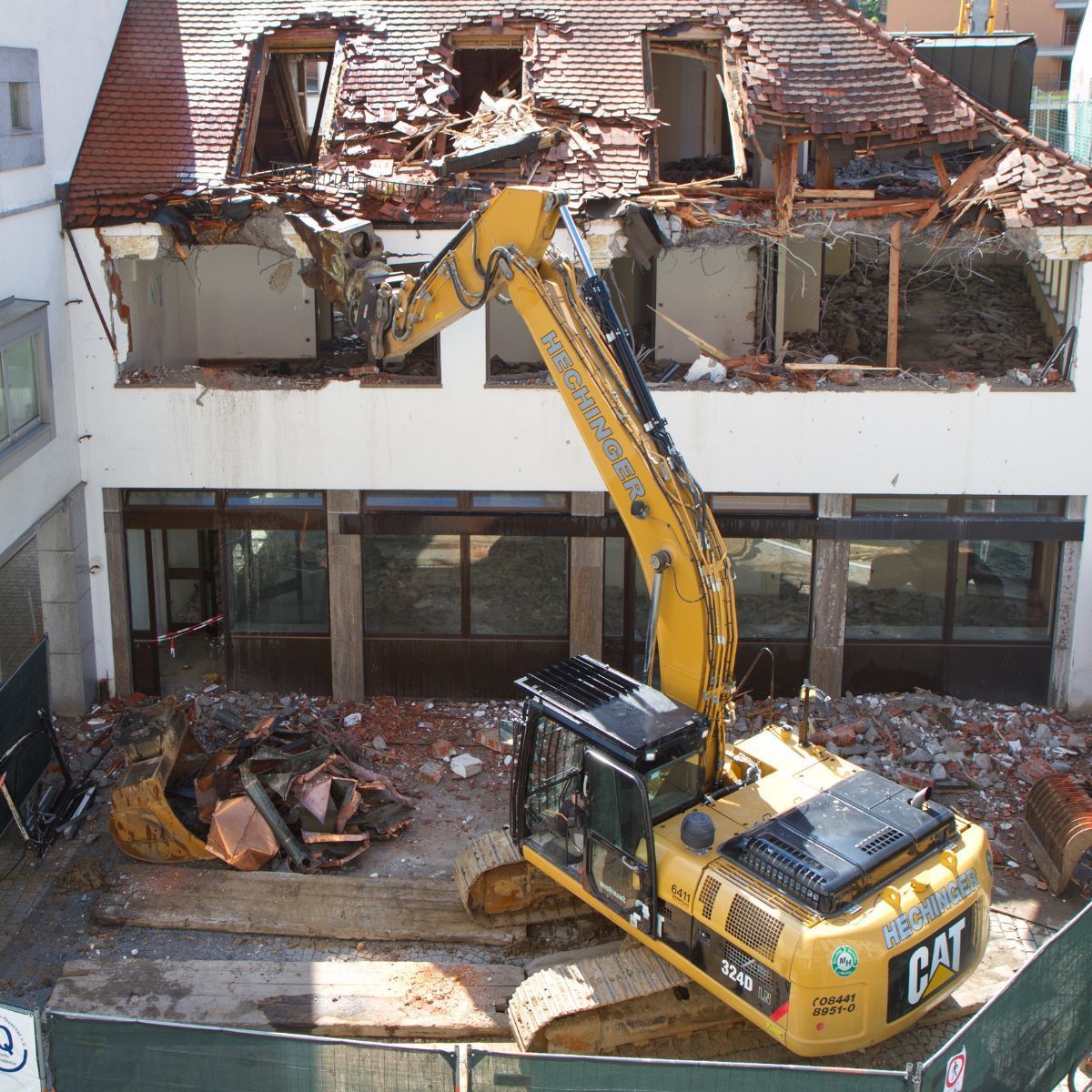 Yellow excavator demolishing a building's top floor; rubble, wood, and broken walls visible.