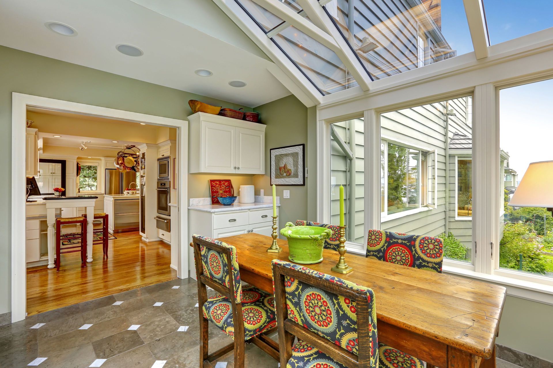 Dining room with glass roof and windows, wooden table and patterned chairs. View into kitchen.