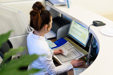 Woman at a reception desk, working on a computer and reviewing paperwork. The desk is white and curved.