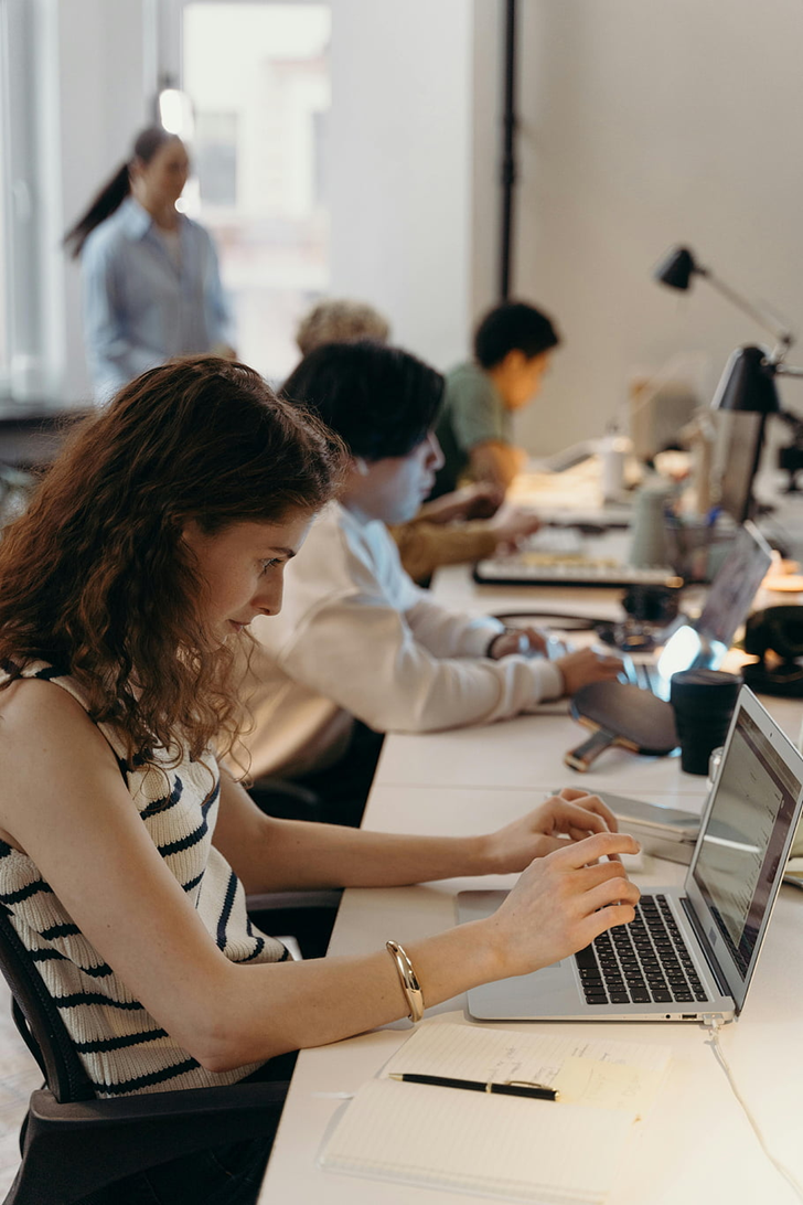 Woman in a striped shirt types on a laptop at a long desk in a bright office, others work in the background.