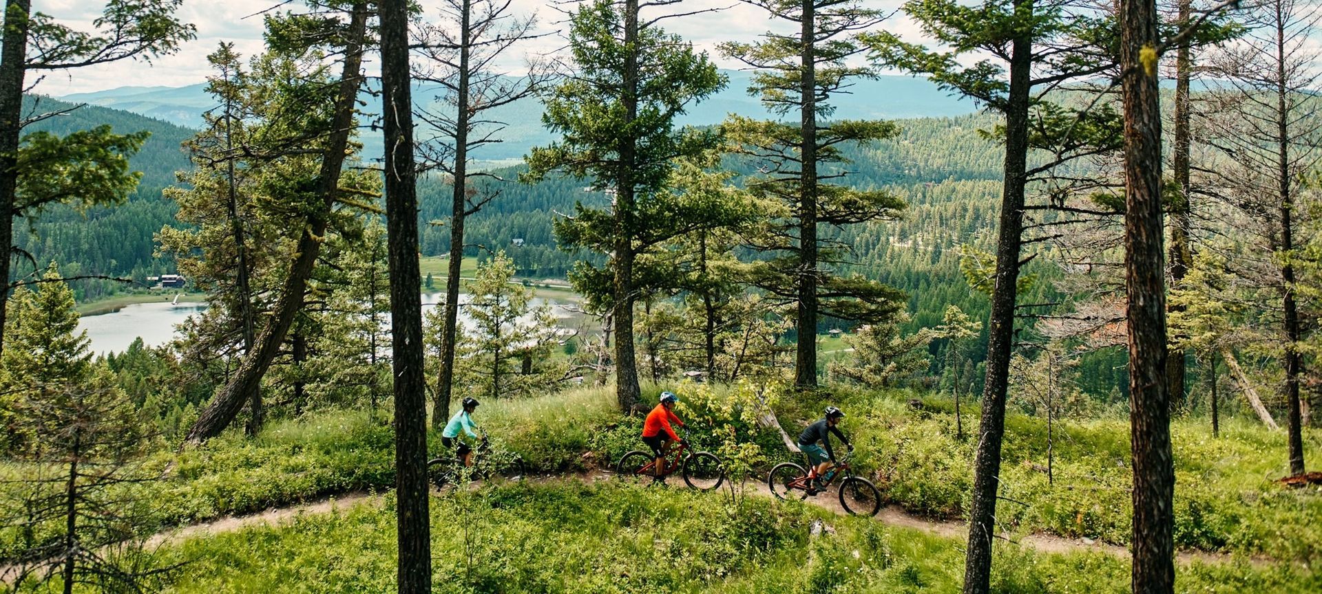 Three cyclists ride mountain bikes on a trail through a forest, overlooking a lake and town.