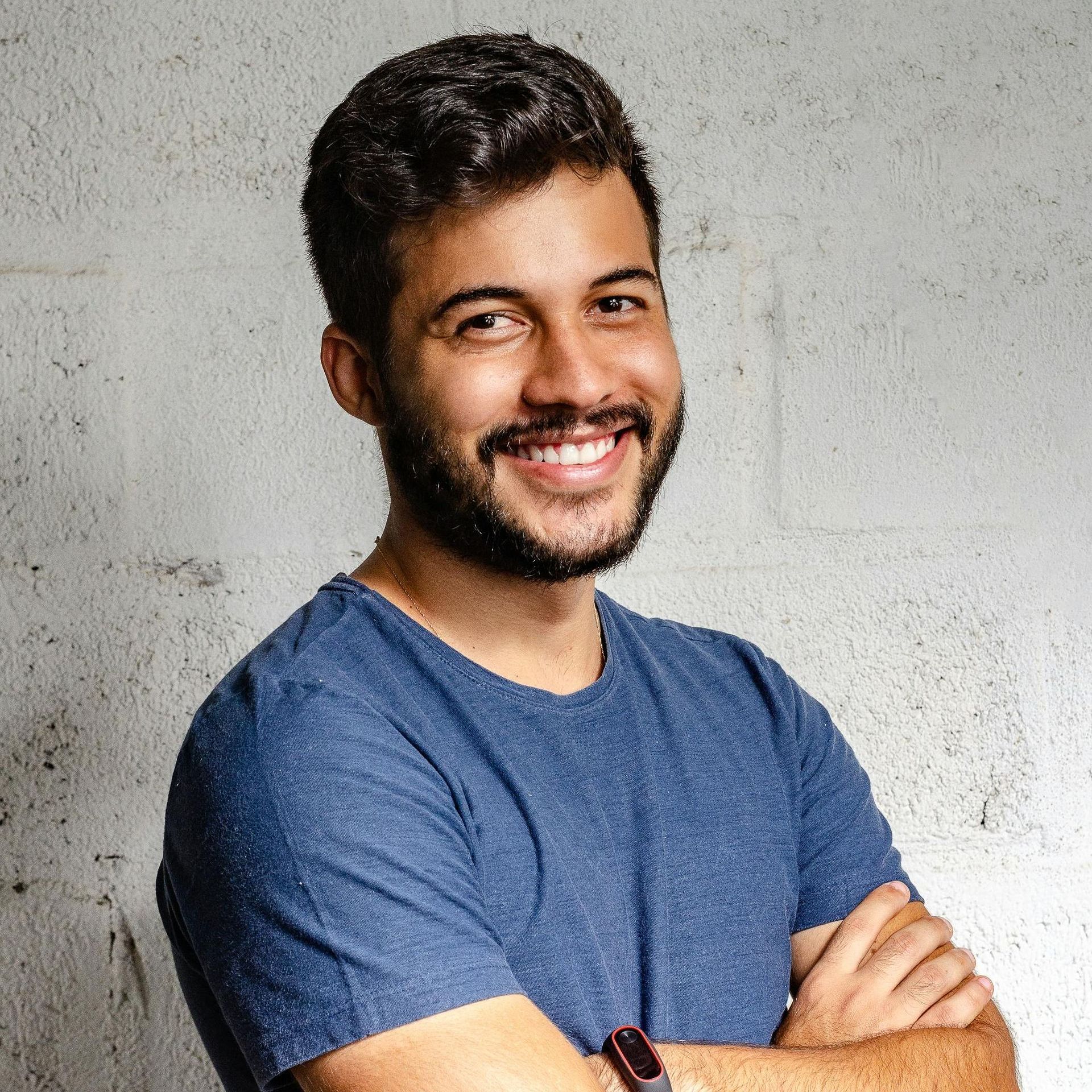 Man with dark hair and beard smiles, arms crossed, wearing a blue shirt, standing against a textured white wall.