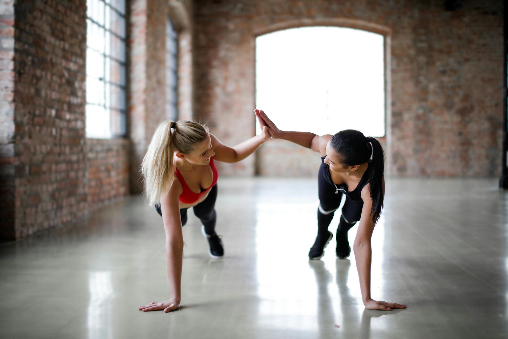 Two women doing push-ups in a studio, one giving the other a high-five.