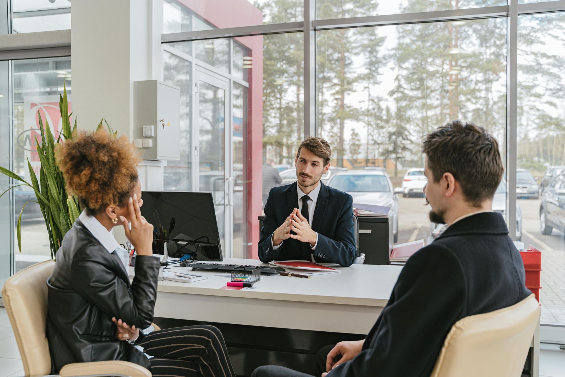 Three people at a desk in an office setting: two clients with a salesperson. Cars are visible outside the large window.