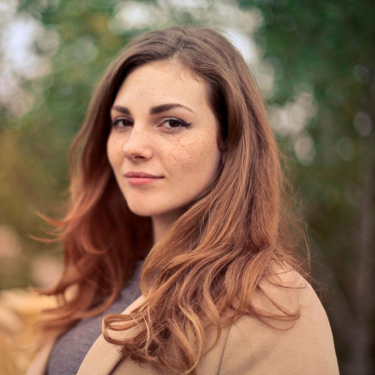 Woman with long auburn hair and freckles, wearing a tan coat, looking towards the camera with a slight smile.