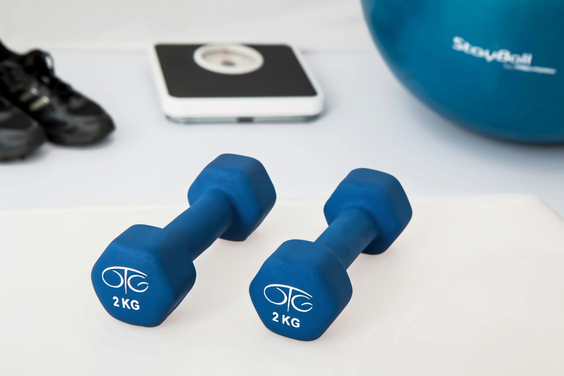 Two blue 2kg dumbbells, scale, shoes, and exercise ball on a white surface.