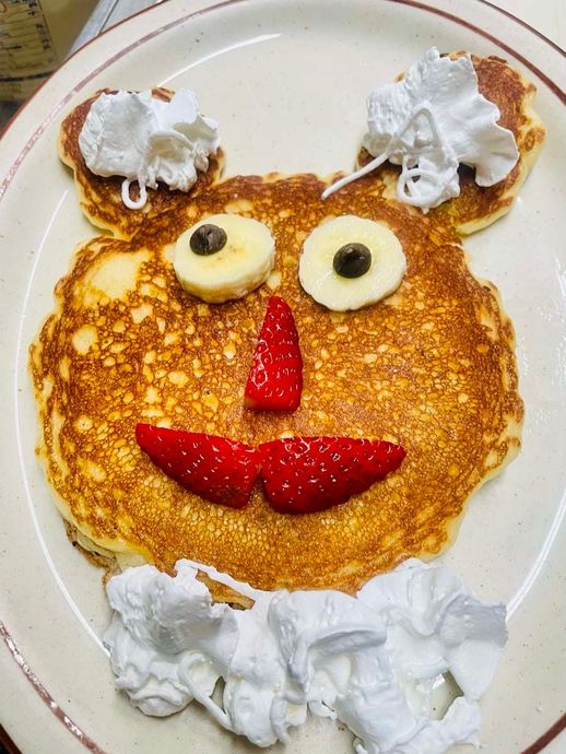 A pancake with whipped cream and strawberries on it is shaped like a teddy bear.