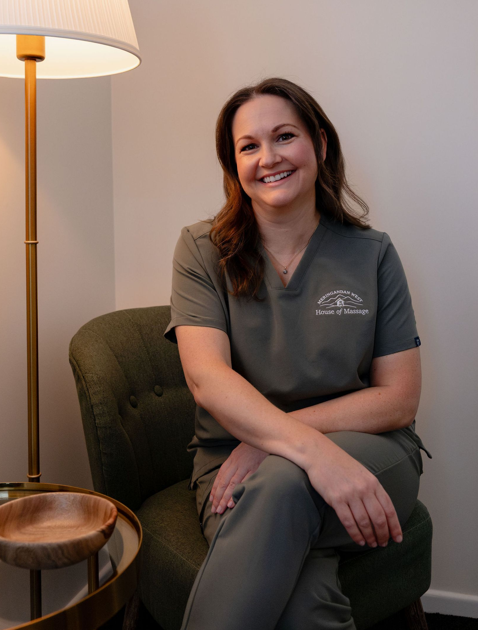 Woman smiling in a green chair beside a lamp and small table, wearing gray scrubs.