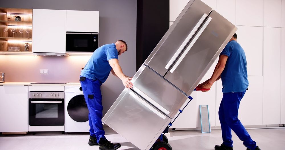 Two men are carrying a refrigerator on a dolly in a kitchen.
