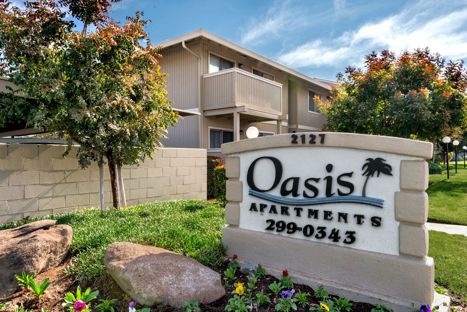 Oasis Apartments sign with a two-story building in the background, trees, and green grass.