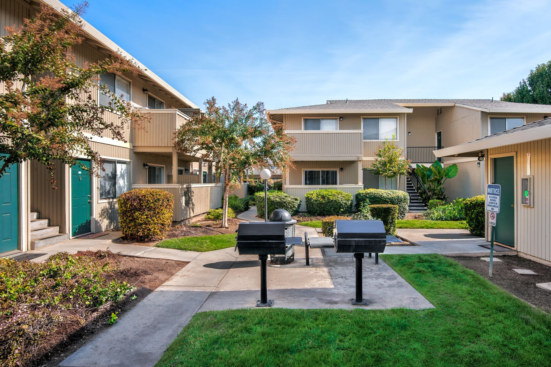 Courtyard with two-story apartments. Grills, tables, and shrubs are in the foreground. Sunny day.