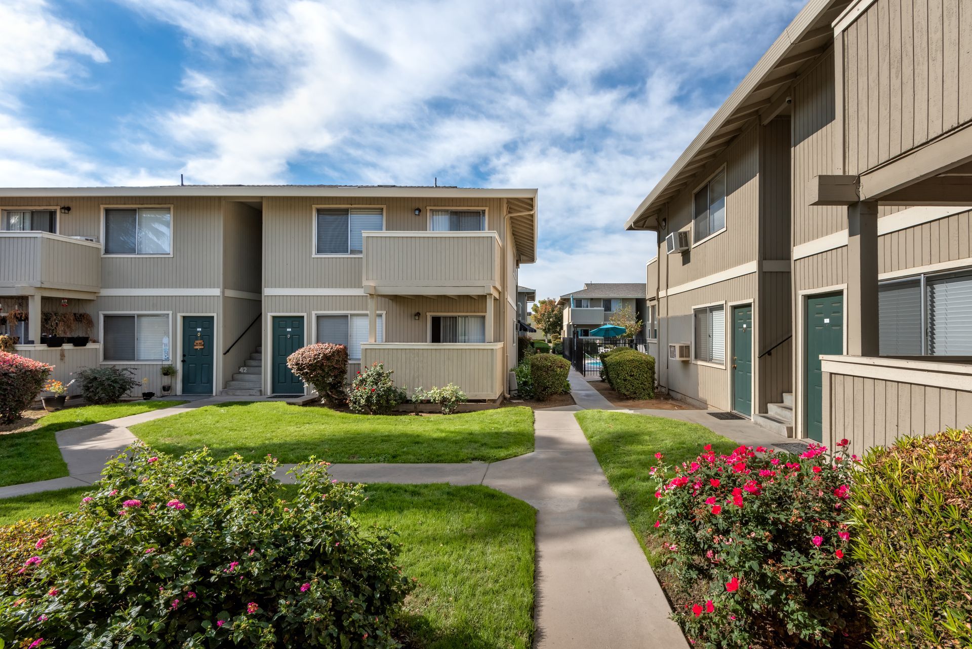 Apartment buildings with beige siding, green doors, and a walkway with green lawns and bushes under a blue sky.