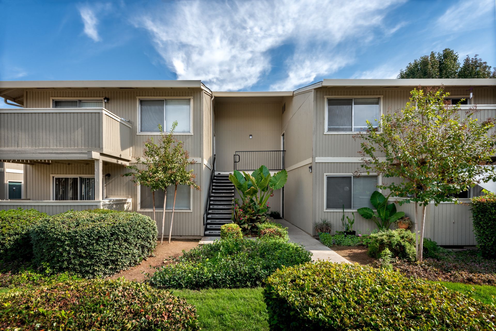 Two-story beige apartment building with stairs, shrubs, and a blue sky.