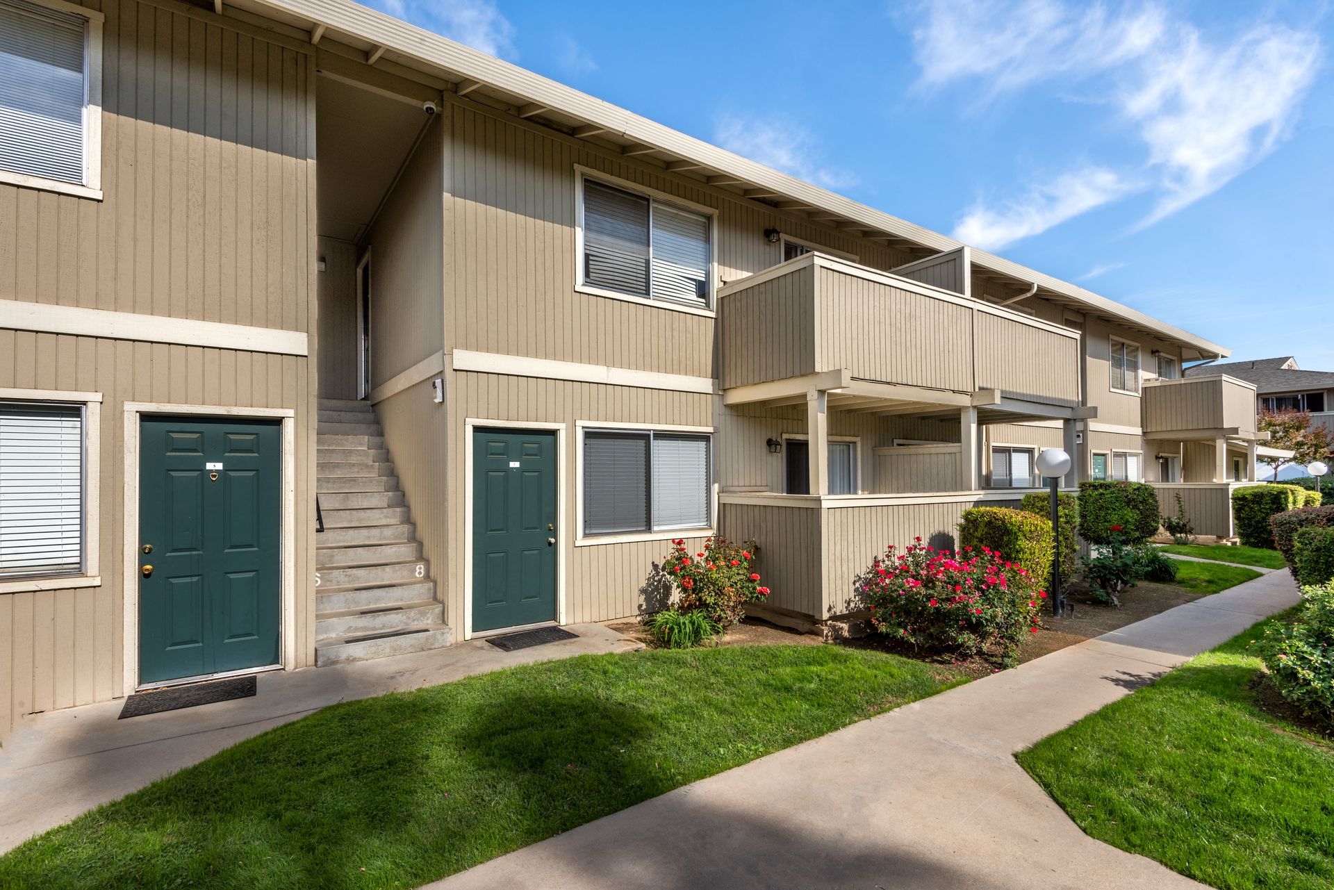 Apartment complex exterior with green doors, beige siding, balconies, and pathway on a sunny day.