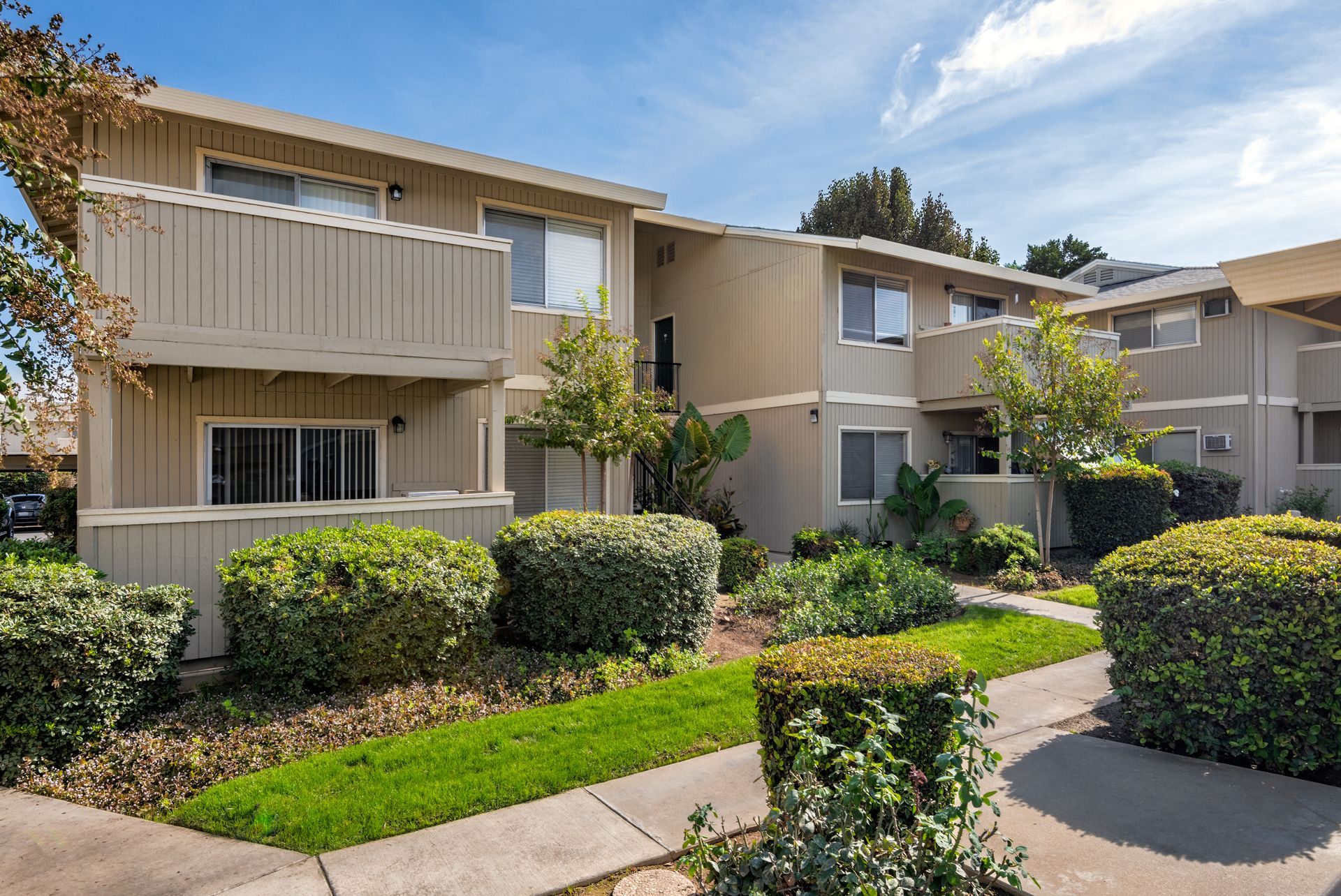 Two-story beige apartment building with small balconies, landscaping, and a clear sky.