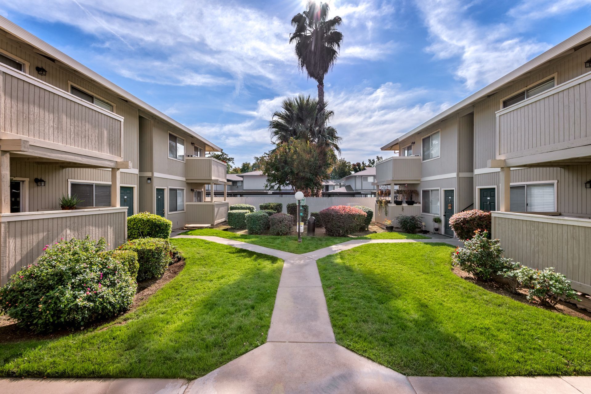 Two-story beige apartment buildings flank a green courtyard with pathways and a tall palm tree under a blue sky.