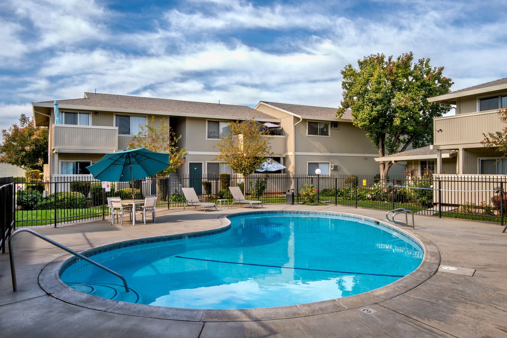 Pool with turquoise water in front of two-story beige apartment buildings; patio table, chairs, and a blue umbrella.