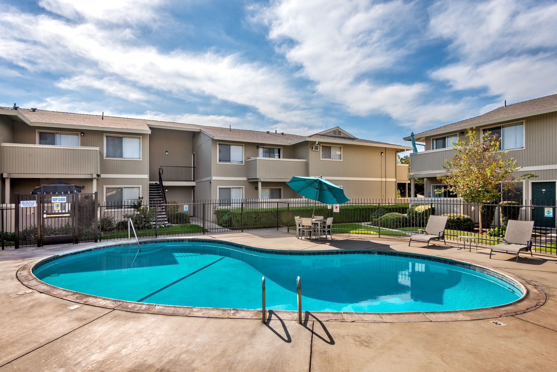 Apartment complex with a pool, umbrella, and outdoor seating under a blue sky.