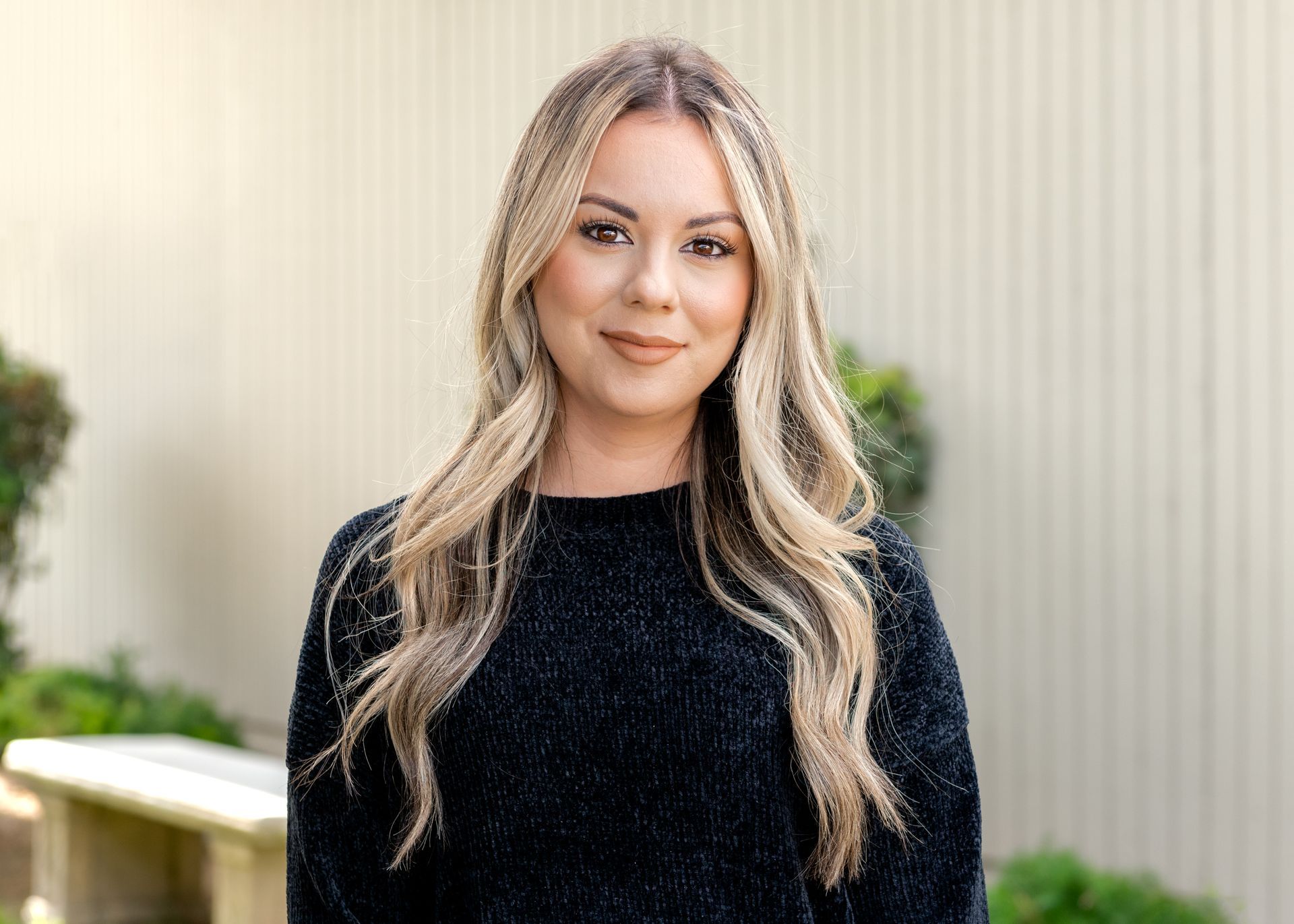 Woman with blonde highlights wearing a black sweater smiles outside against a cream-colored wall.