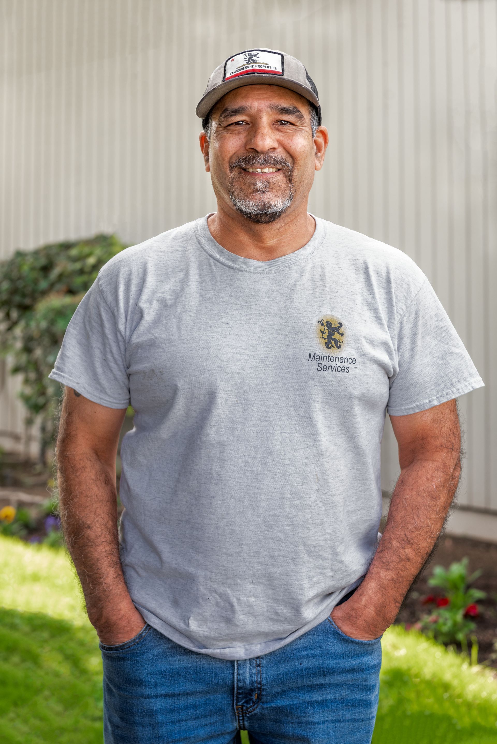 Man in cap, gray shirt, and jeans with hands in pockets, standing in front of a building and grass.