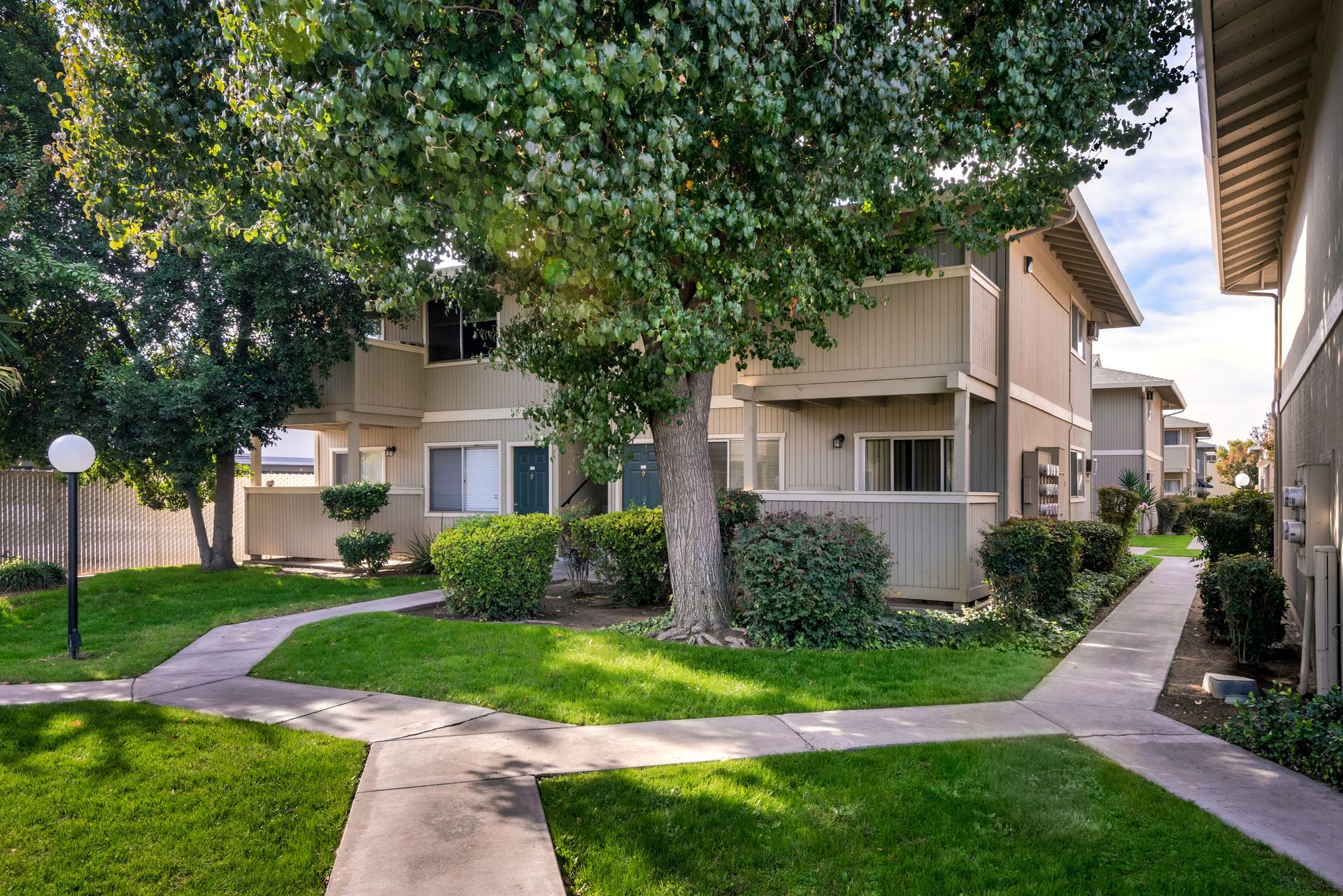 Apartment complex with green lawns, pathways, trees. Beige buildings, blue sky.