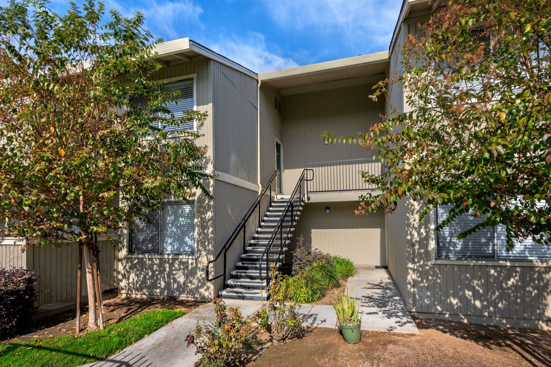 Two-story beige apartment building with black staircase, green plants, and blue sky.