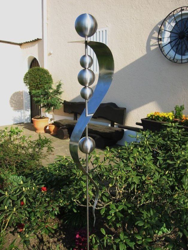 Eine Edelstahlskulptur in einem Garten mit einer Uhr im Hintergrund