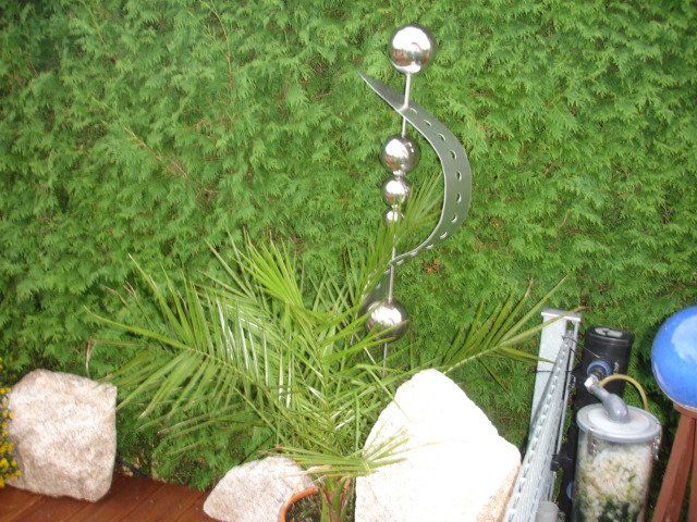 Eine Metallskulptur steht auf einem Holztisch neben einer Pflanze und Felsen.