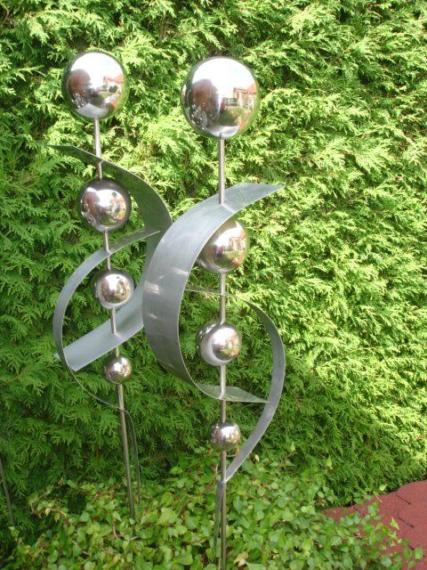 Eine Edelstahlskulptur sitzt vor einem Busch.