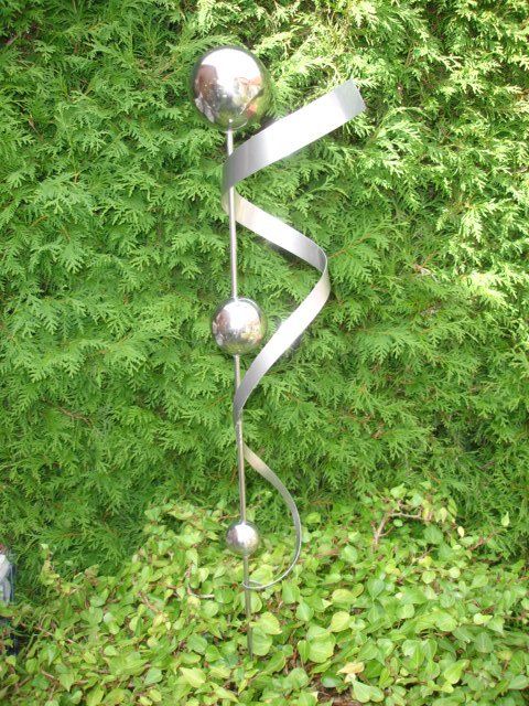 Eine Edelstahlskulptur sitzt vor einem Busch.