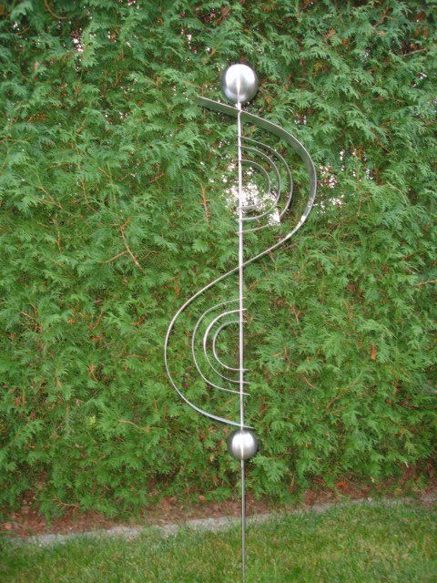 Eine Edelstahlskulptur sitzt im Gras vor einer Hecke.