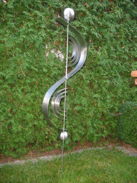 Eine Edelstahlskulptur sitzt im Gras vor einer Hecke.