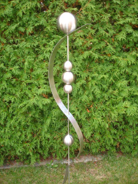 Eine Edelstahlskulptur sitzt im Gras vor einem Busch.